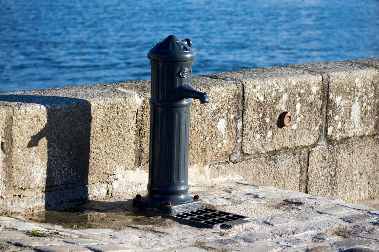 A classic water pump by a seaside stone wall, illuminated by sunlight, with a serene blue sea backdrop.